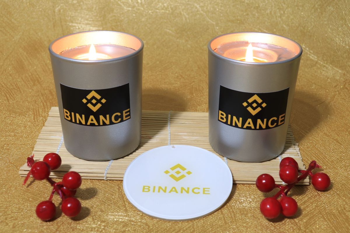 Two silver candles with Binance labels on a bamboo mat, gold background and red berries