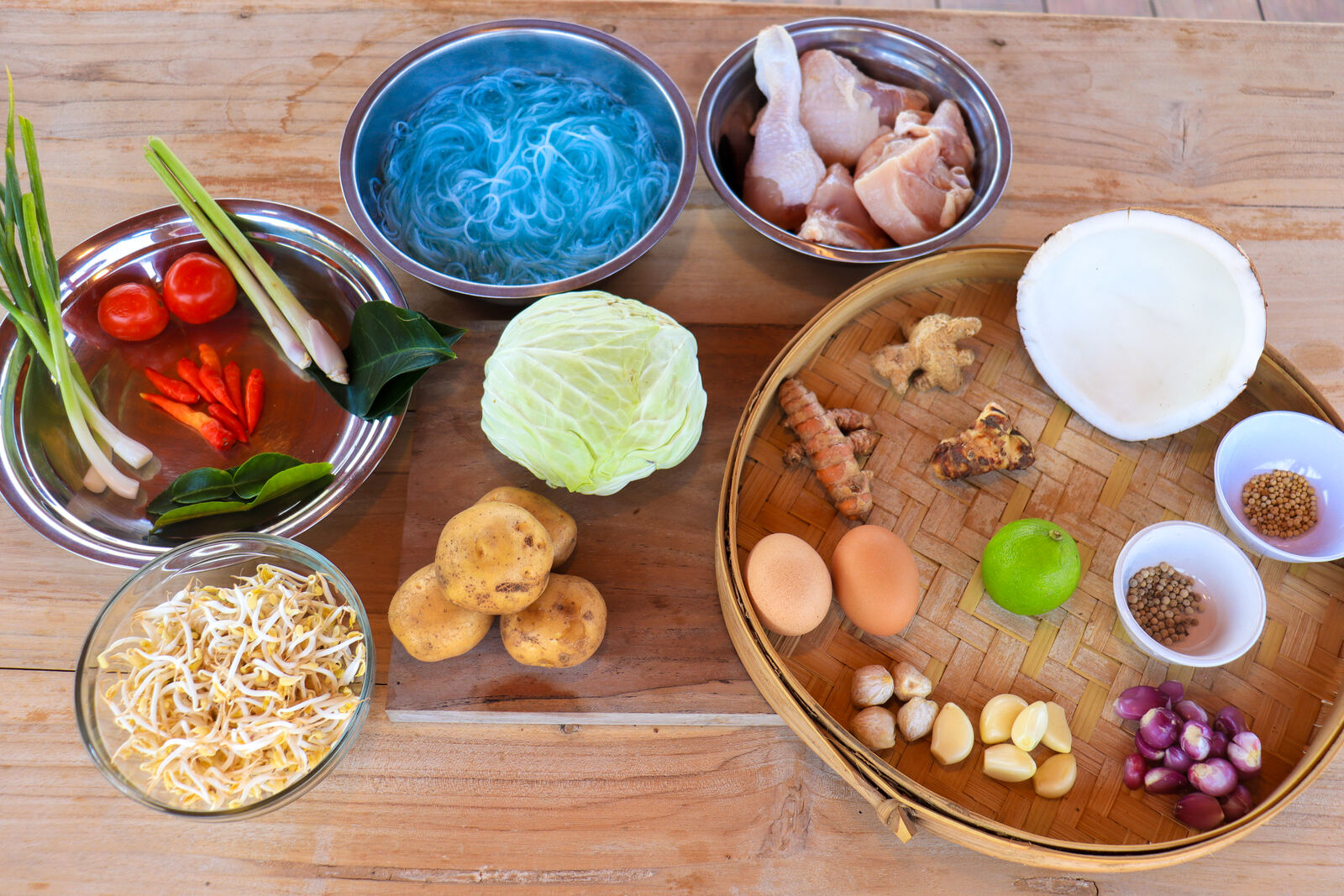 Fresh ingredients for traditional Indonesian cooking laid out on a wooden table: spices, vegetables, chicken and noodles