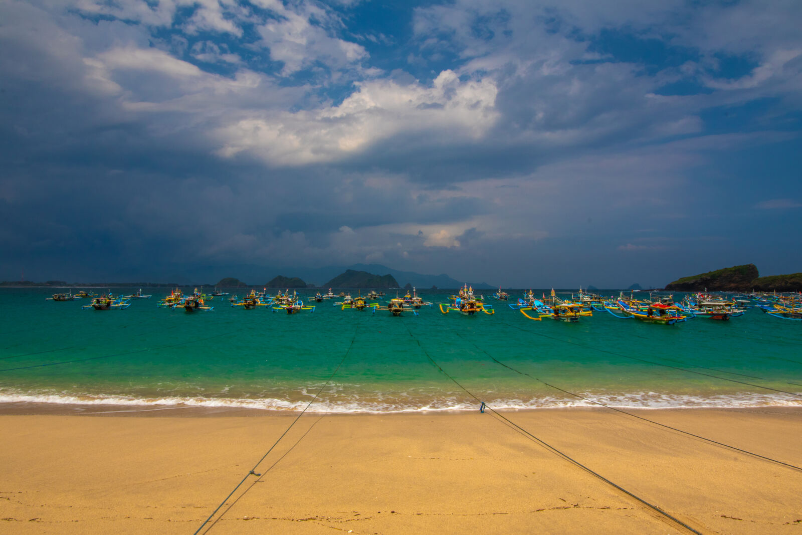 Turquoise bay with traditional colorful fishing boats under a stormy sky, seen from a sandy beach