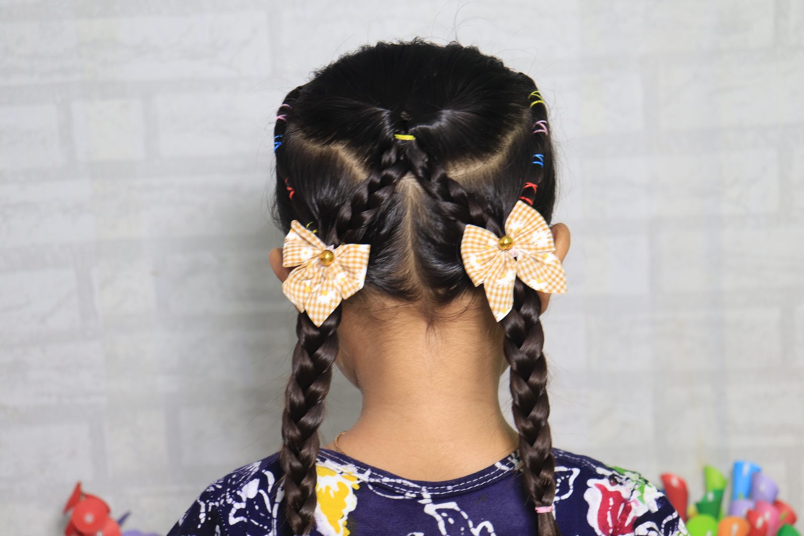 Back view of a girl with two braids and orange gingham daisy hair bows against a light wall
