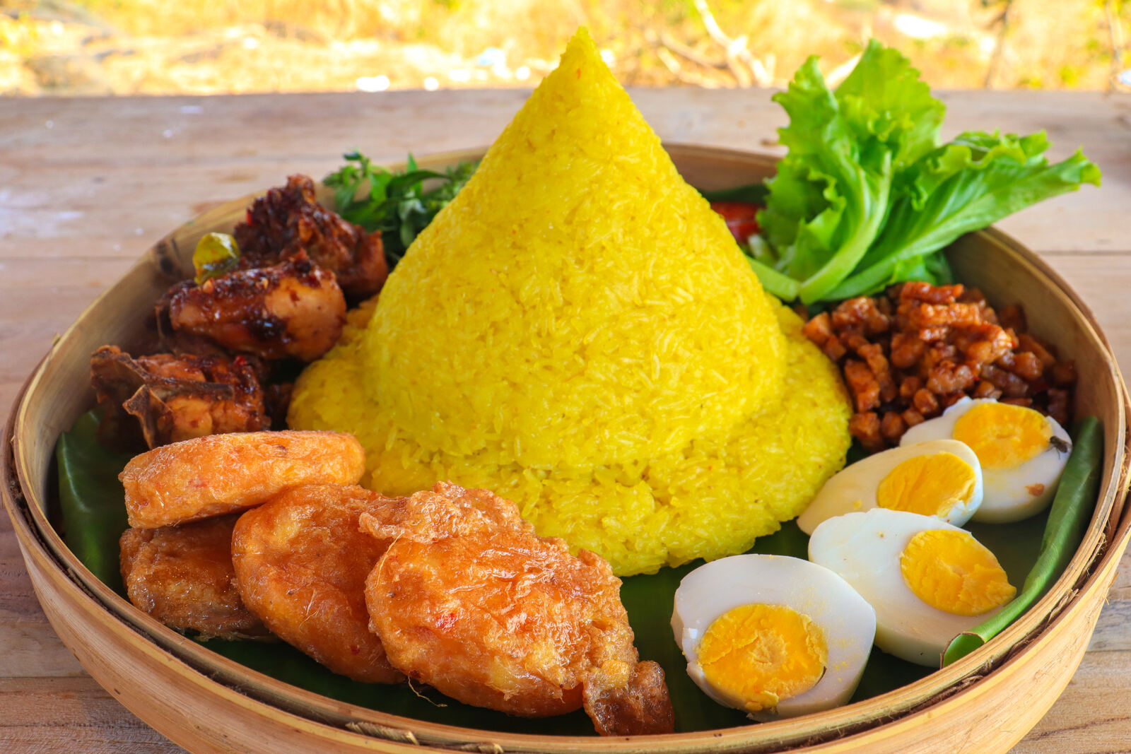 Yellow rice tumpeng with fried side dishes and boiled eggs on a bamboo tray