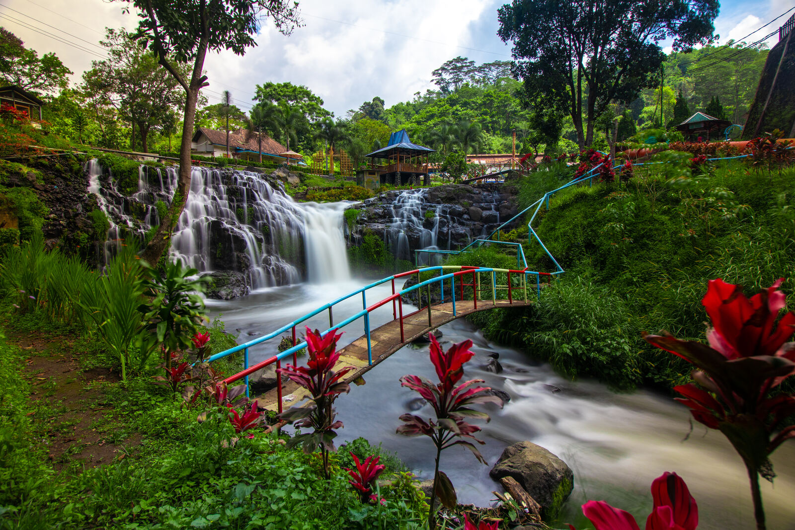 Waterfall and a small colorful footbridge in a lush green park with red tropical plants
